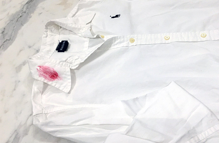 A white shirt laid out on a marble countertop with a red lipstick stain on the collar about to be laundered with The Laundress method for removing lipstick stains.